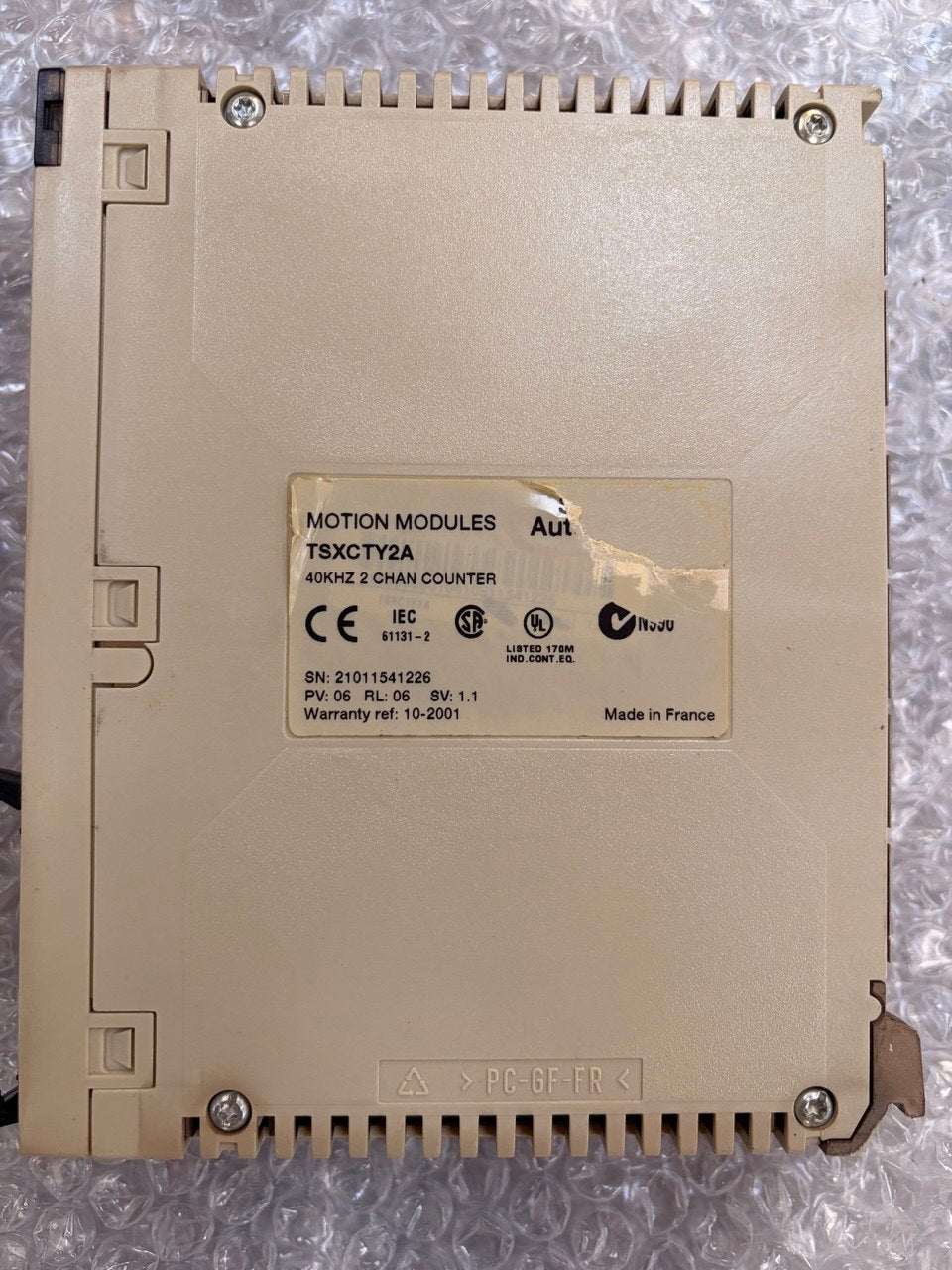 A beige industrial motion control module labeled TSXOTY2A, photographed from above on a crinkled bubble wrap surface. The back shows screw terminals, ribbed edges, and a central specification sticker.