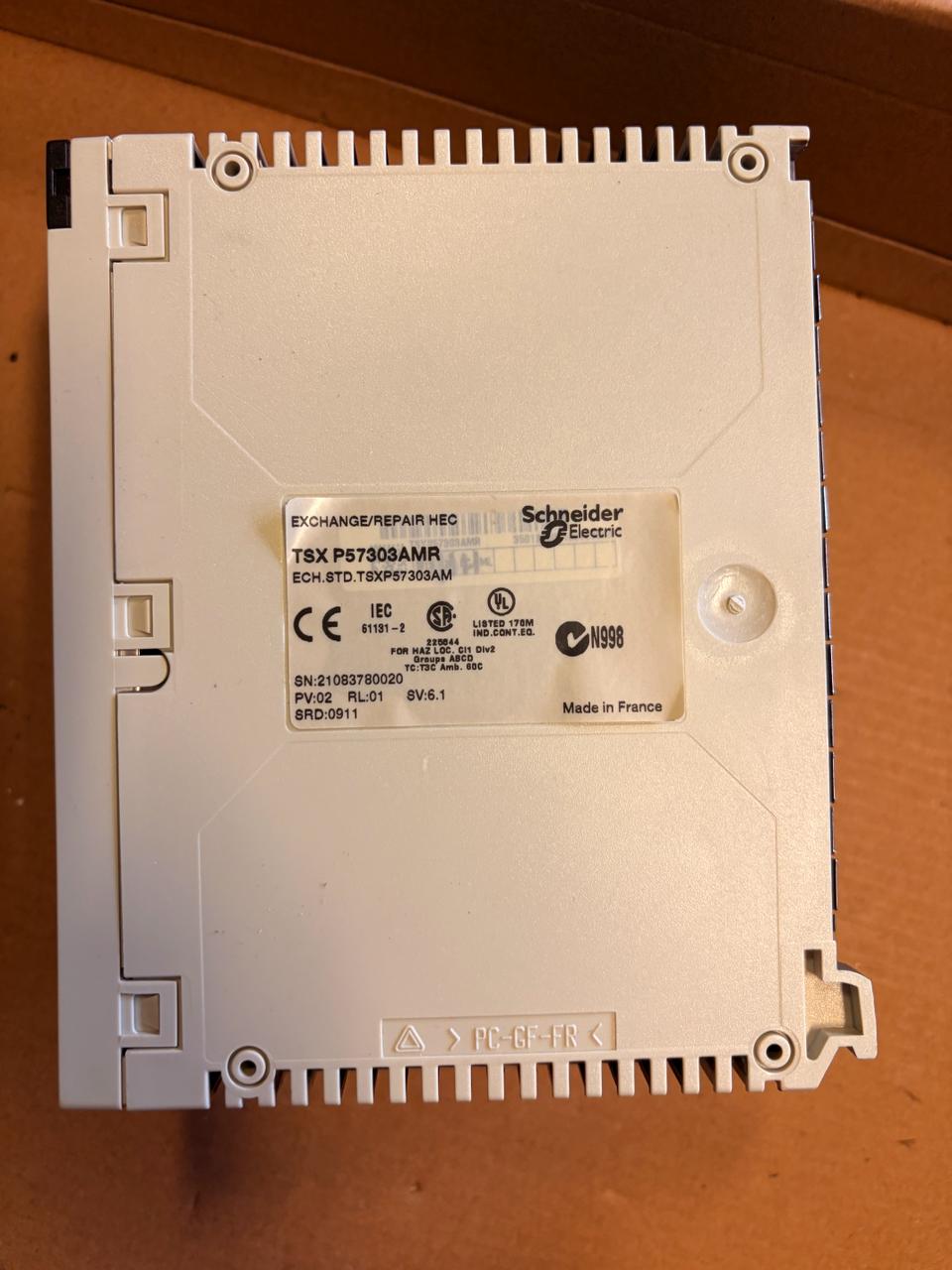 A Schneider Electric TSX P57303AMR industrial electronic module, showing the back side with a white plastic housing and a central label, placed on a brown cardboard surface.