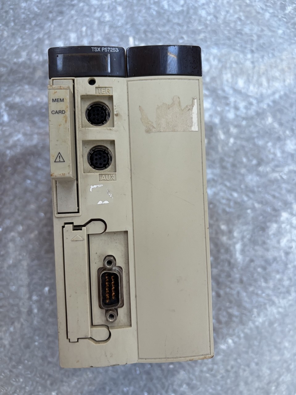 A vintage industrial electronic module, specifically a Mitsubishi TSX PF28S unit, shown from the rear. The beige plastic housing shows signs of wear, scuffs, and a sticker residue on the front panel, with dark gray/black connectors and a VGA port visible.